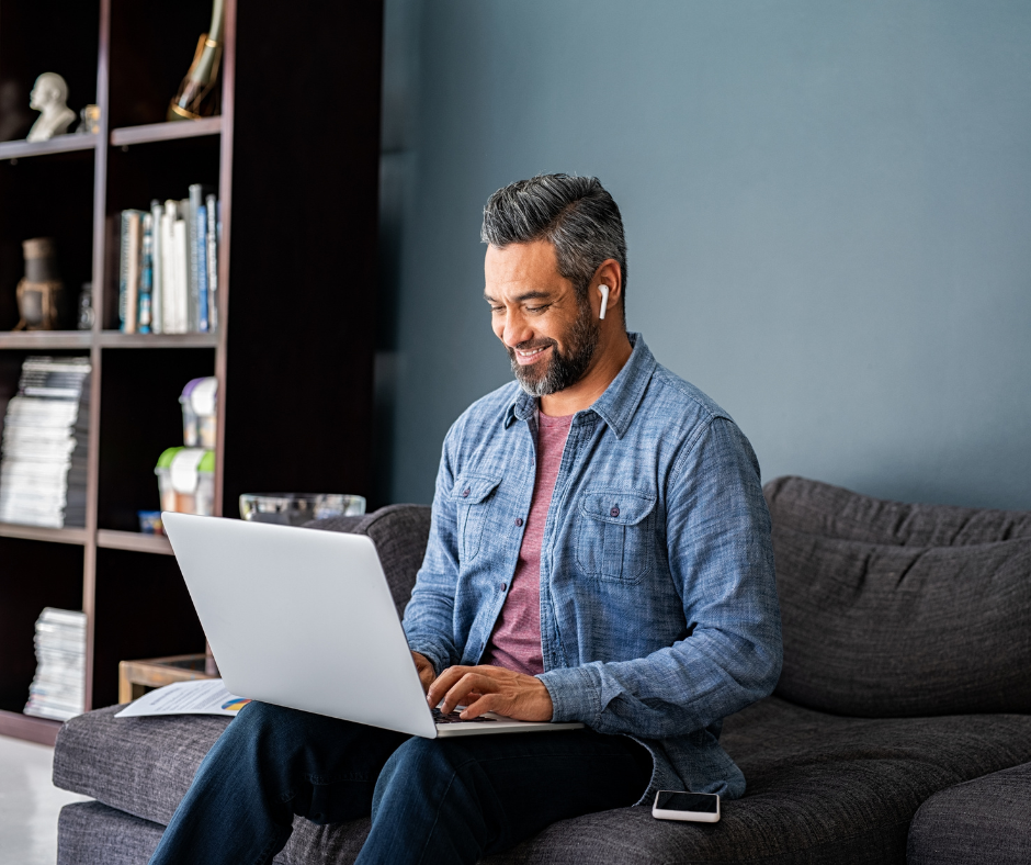 Mature Indian male sitting on couch typing on laptop with wirless earphones in. 