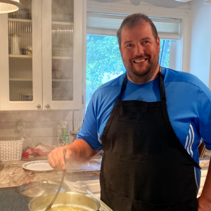 Photo of Dave Carragher standing and smiling in his kitchen stirring a pot of noodles.