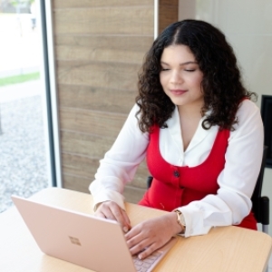 Photo of Kerry wearing a red dress sitting at a desk and working on a laptop computer.